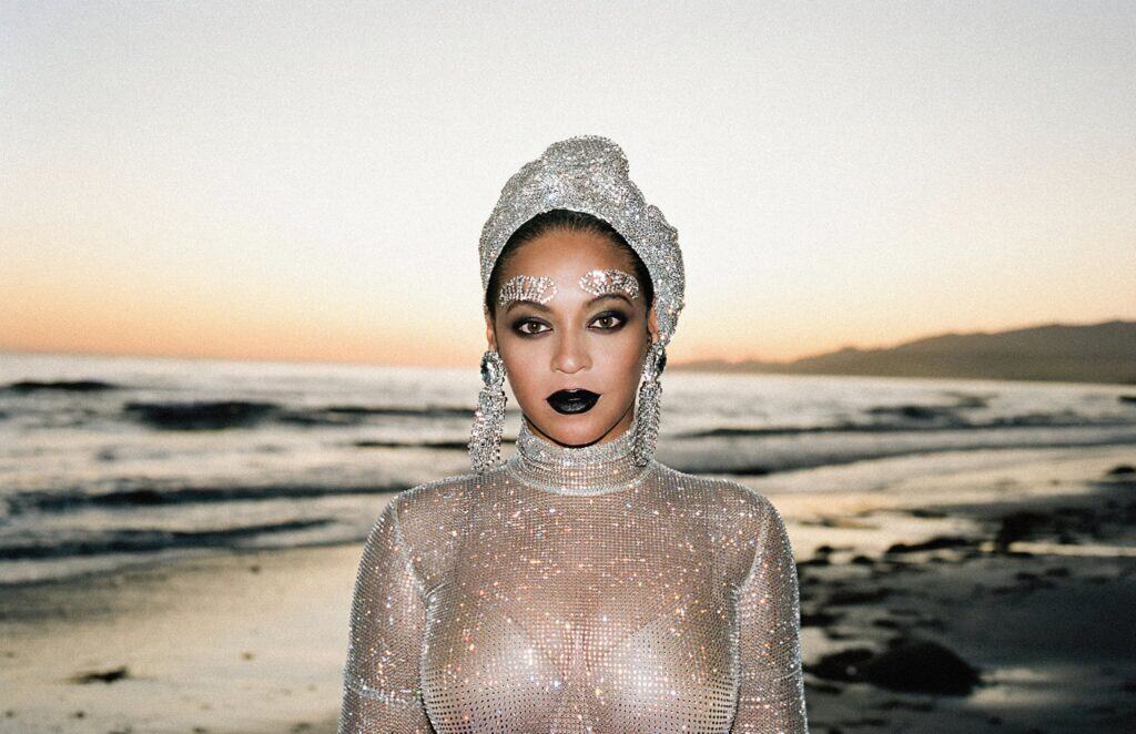 Beyoncé, mulher negra, em vestimenta furta-cor e em fundo de praia e pôr do sol
